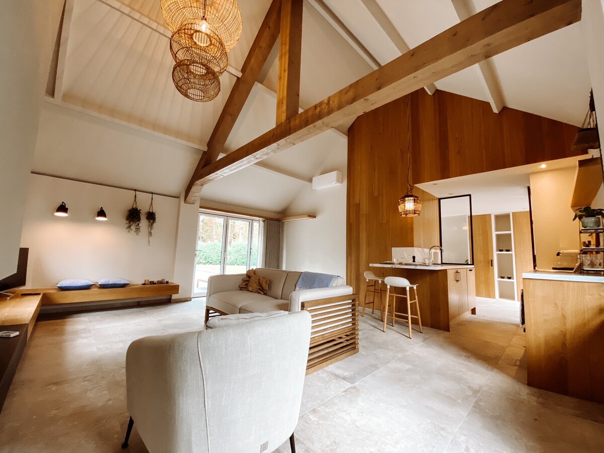 An open living area showcases a high ceiling supported by wooden beams, with a comfortable seating arrangement. A cozy nook is positioned by large windows, allowing natural light to fill the space. A modern kitchen is visible, featuring wooden cabinetry and seating at the bar.