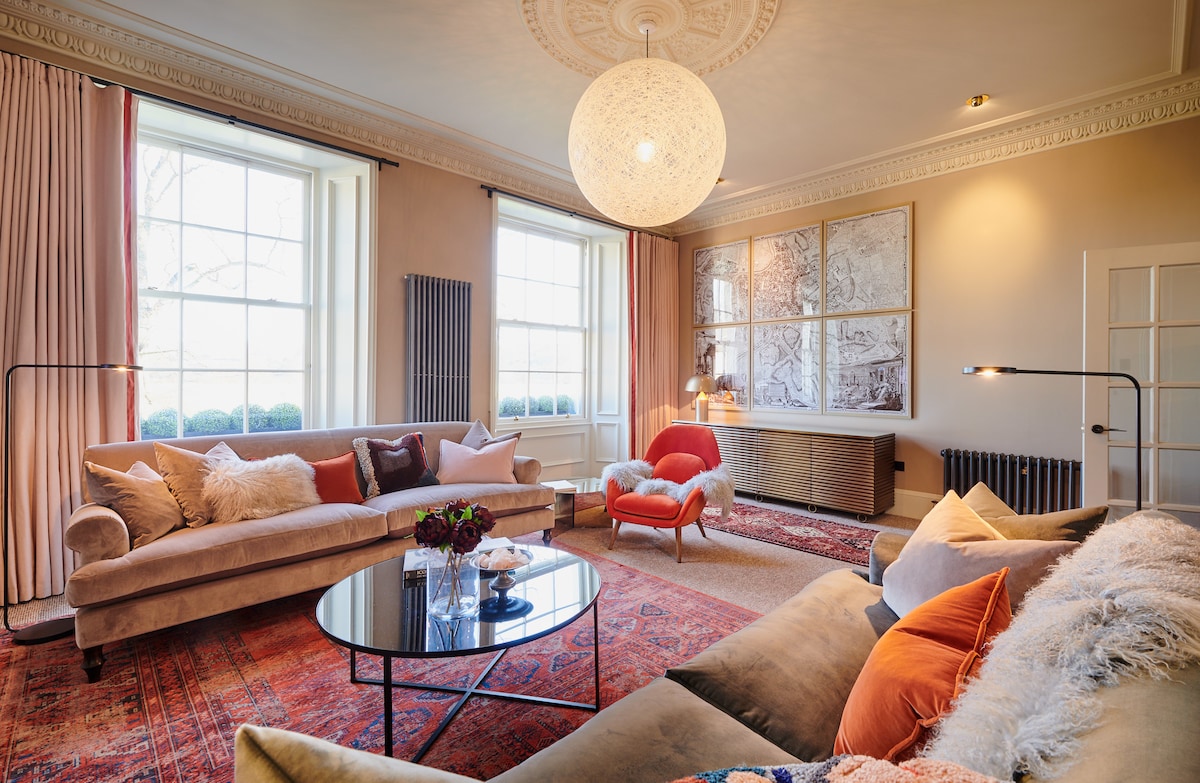 A spacious living area features high ceilings and large windows allowing natural light to illuminate the room. A soft couch and armchair in warm tones are paired with a glass coffee table, while decorative artwork accents the walls. A textured rug adds warmth beneath the seating arrangement.