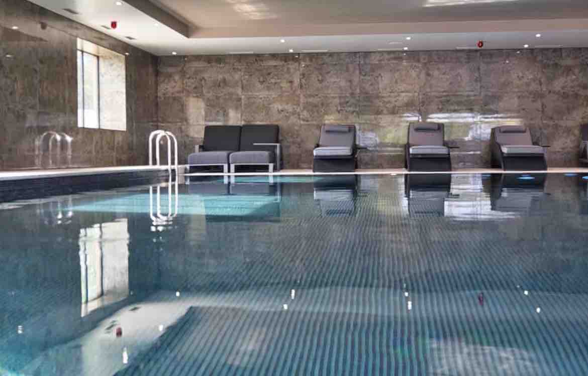 A serene indoor swimming pool features a glossy surface reflecting the ambient lighting. Lounge chairs are positioned along the edge, and contemporary stone walls provide a calming backdrop. The atmosphere conveys a sense of tranquility and relaxation.