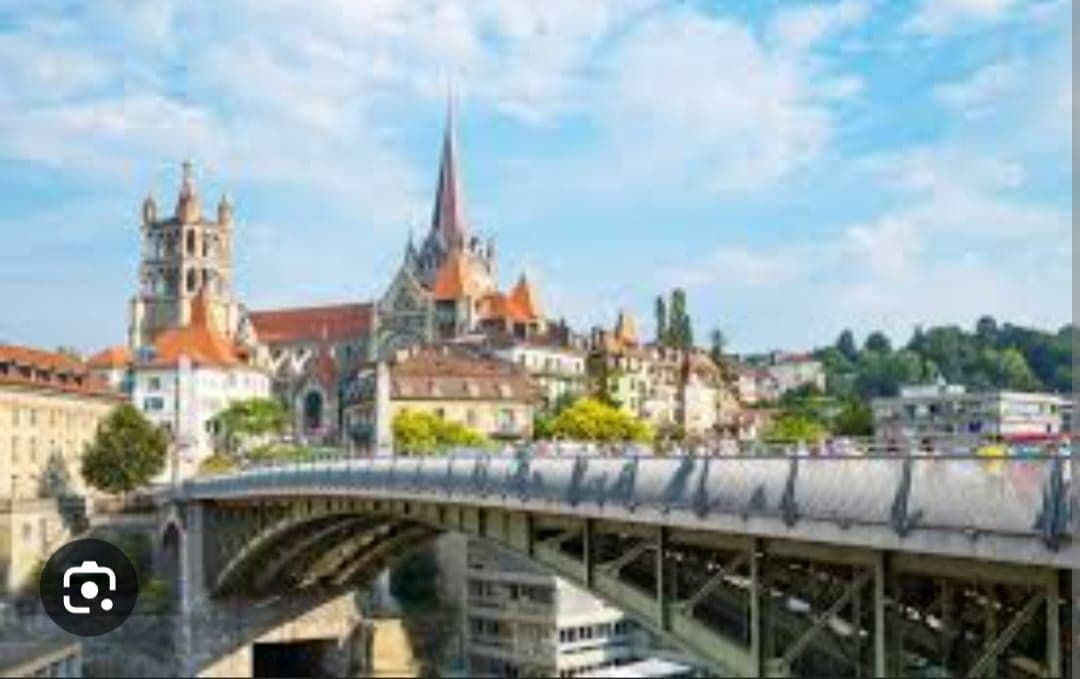 A cityscape is visible, featuring a bridge that spans a river. Historic buildings with distinct architectural styles and colorful roofs line the riverbank. A prominent church spire rises in the background among green trees and a blue sky adorned with scattered clouds.