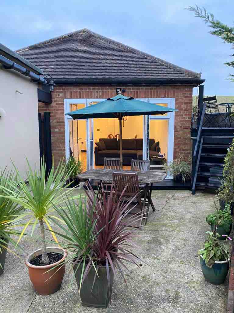 A private outdoor dining area is showcased, featuring a wooden table with six chairs under a green umbrella. Surrounding potted plants, including spiky palm varieties and greenery, enhance the space. The background reveals a large glass door leading into a cozy living area.