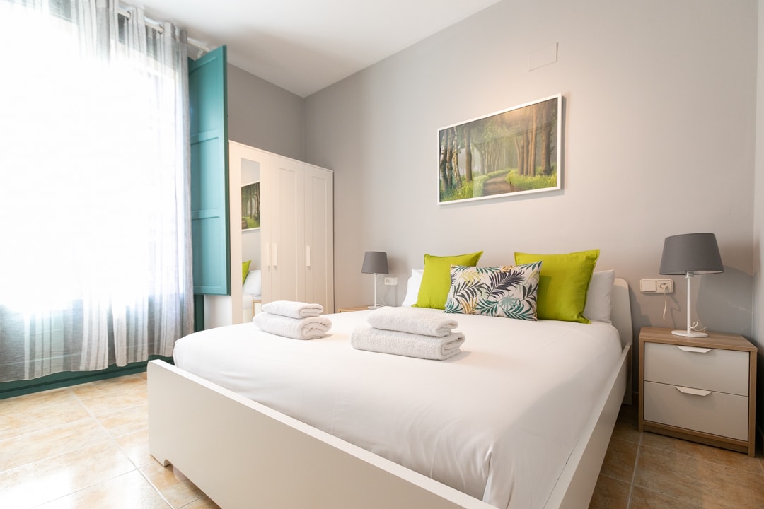 A bright sleeping area is equipped with a spacious bed, adorned with neatly arranged towels. A large window with sheer curtains allows natural light to fill the room. A light-colored wardrobe and bedside tables provide functional storage, complemented by framed artwork depicting a serene forest scene.