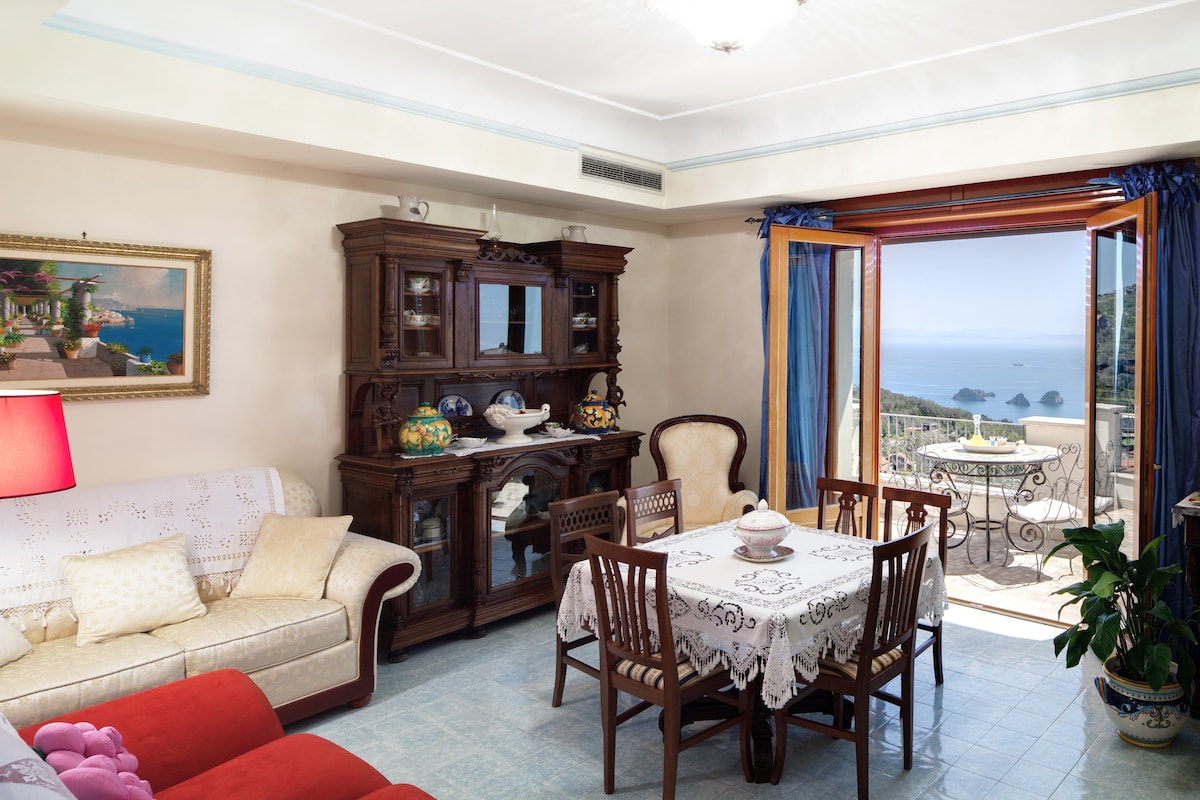 A welcoming living area displays a mix of classic and contemporary furnishings. A comfortable sofa and elegant dining table are set against large windows that offer ocean views. Decorated with tasteful art and plants, the space creates an inviting atmosphere for relaxation.