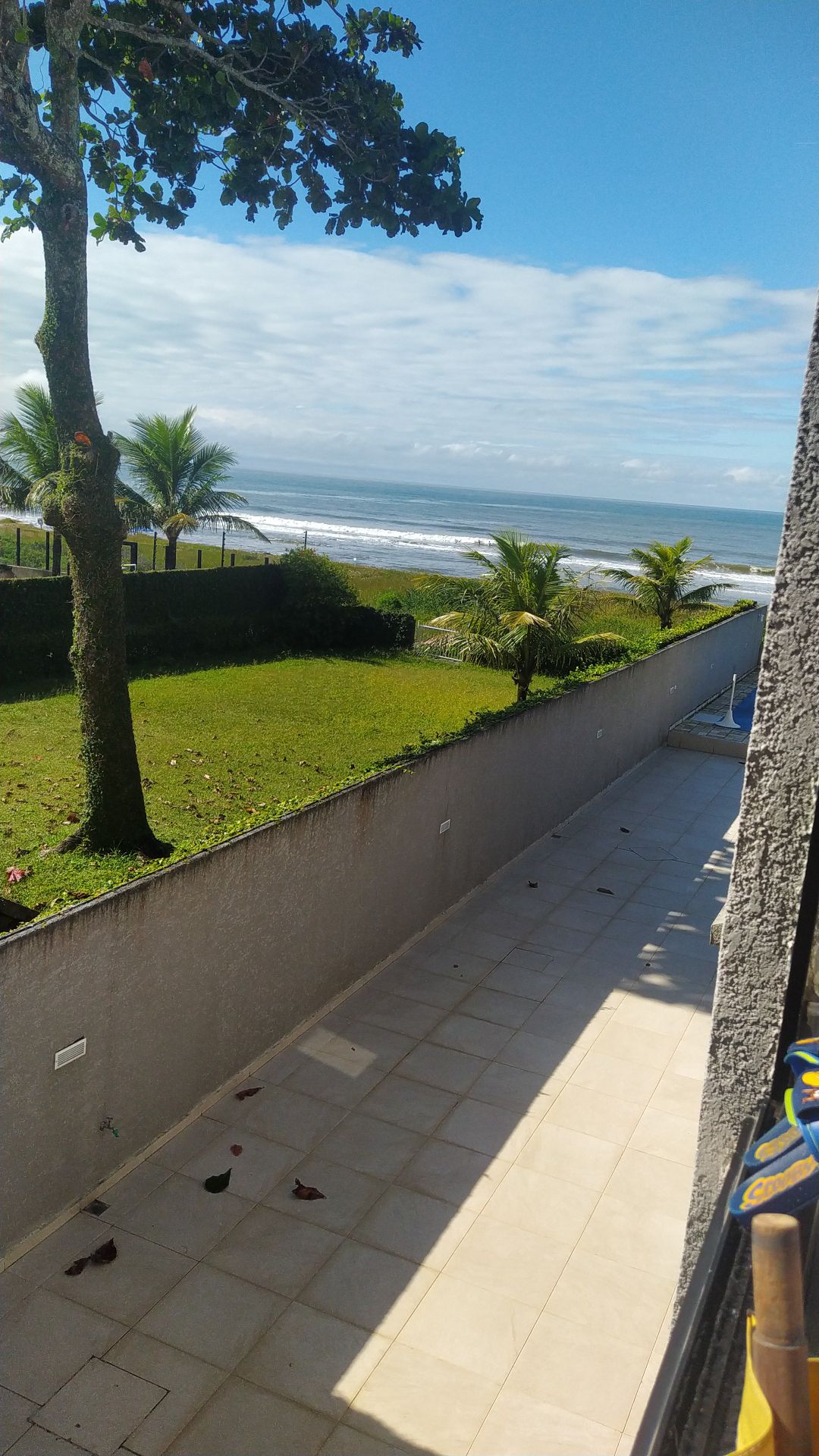 An outdoor view captures the landscaped garden leading towards the beach. Lush palm trees are visible, and the ocean can be seen in the distance, framed by the clear blue sky. The tiled path extends along the property, providing access to the beach.