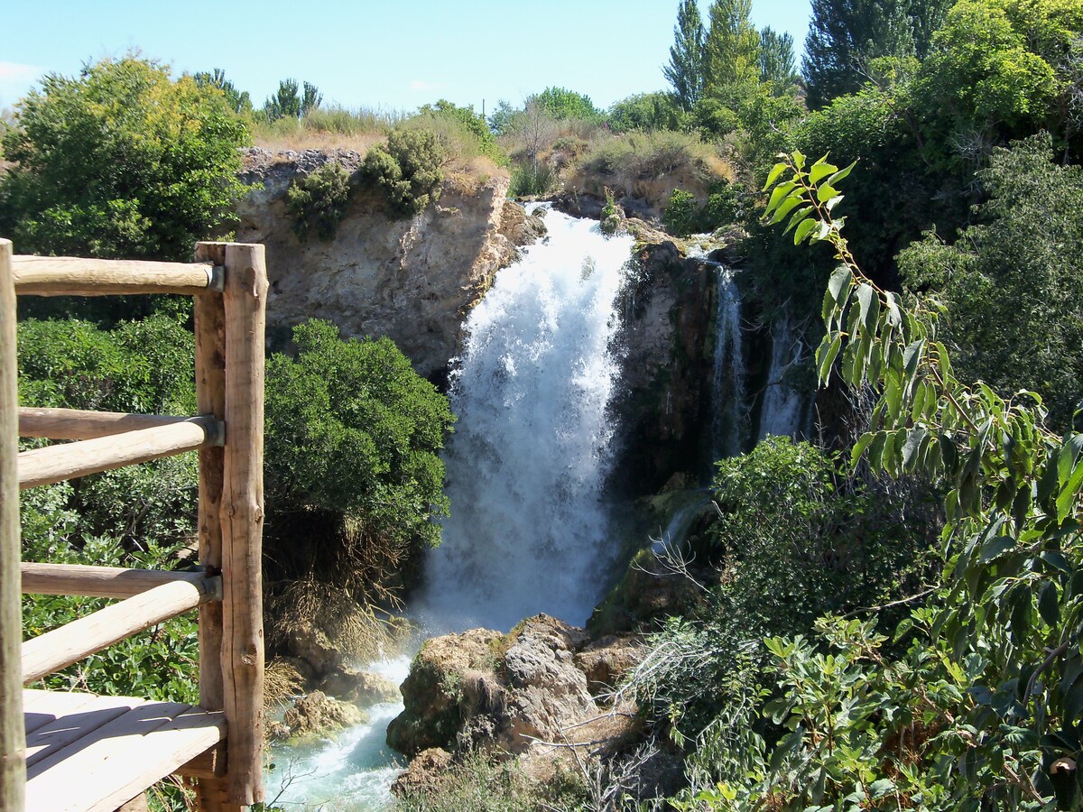 A cascading waterfall is surrounded by lush greenery, with the water flowing down rocky ledges. The vibrant foliage contributes to the natural setting, evoking a sense of tranquility. A wooden railing is visible in the foreground, providing an inviting viewpoint to admire the scenery.