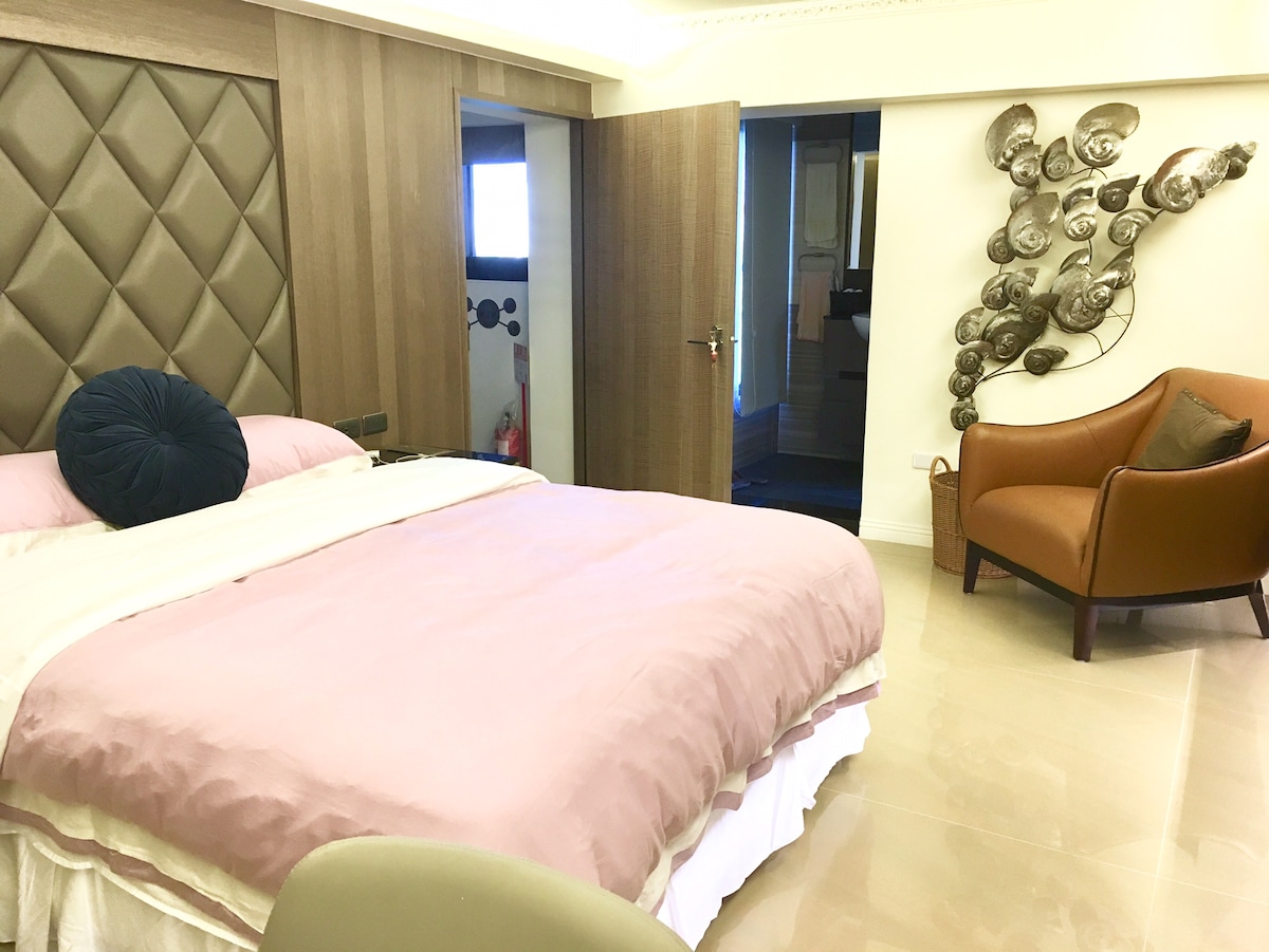 A well-appointed bedroom features a spacious bed with soft, pastel bedding and a decorative pillow. A modern armchair complements the elegant decor. The textured wall behind the bed and artistic wall sculpture enhance the inviting ambiance of the room.