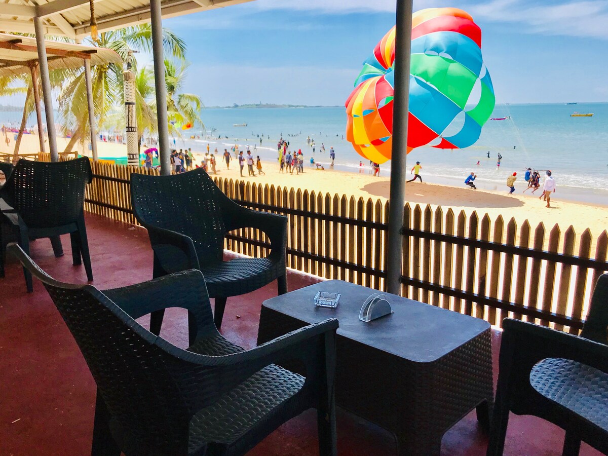 A covered balcony features dark wicker chairs and a small table, offering a view of the beach below. Colorful parasailing can be seen above the water, with people enjoying beach activities and swaying palm trees lining the shore.