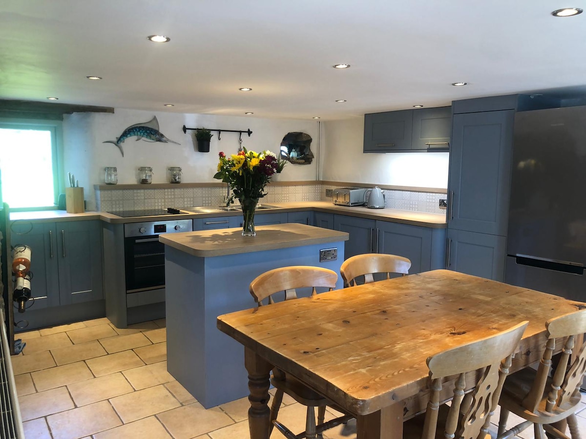 A spacious kitchen is equipped with modern appliances and a large central island. A wooden dining table with six chairs is placed in the center. Natural light enters through a window, enhancing the welcoming environment. Decorative wall art adds a nautical touch.