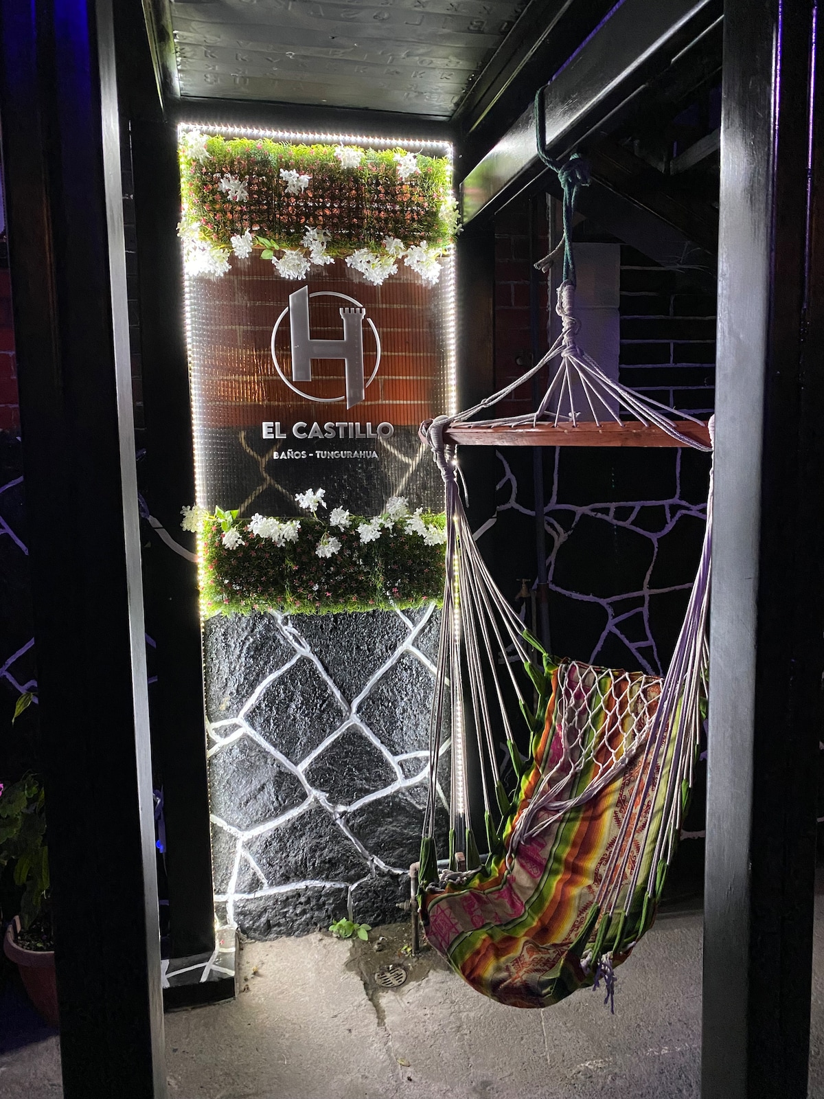 A welcoming entrance features a vibrant hanging hammock, framed by decorative plants and illuminated signage displaying the name 'El Castillo.' The exterior is complemented by a textured stone wall, enhancing the overall ambiance of relaxation and comfort.