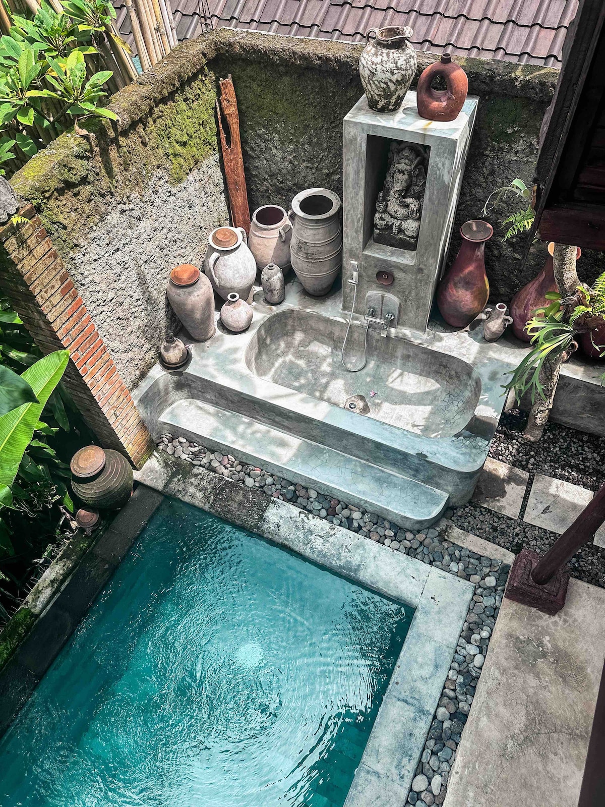 An outdoor bathing area features a stone bathtub and a serene swimming pool. Surrounding the space are various intricately designed earthenware pots set against a textured wall, enhancing the tranquil atmosphere with greenery visible in the background.