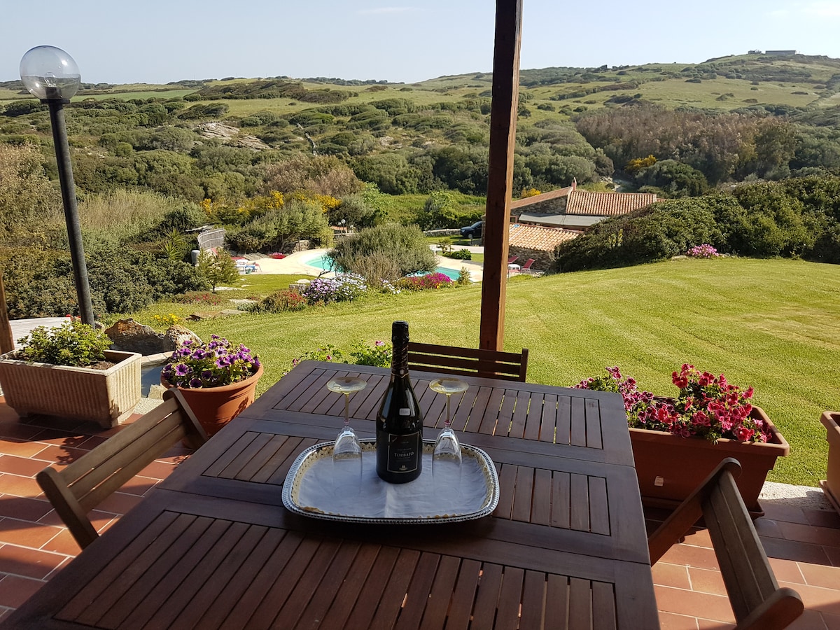An outdoor dining area features a solid wooden table surrounded by several chairs. A tray with glasses and a bottle of wine is placed center stage. In the background, lush greenery and colorful flowers lead to a swimming pool, set against gentle hills under a clear sky.