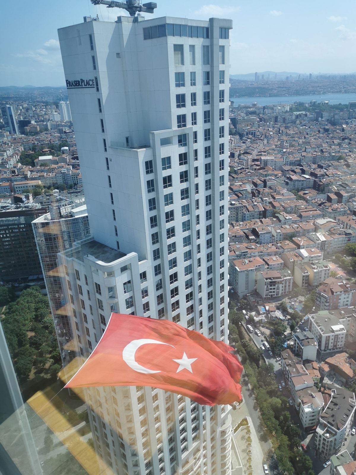 A stunning aerial view captures a towering building against a clear blue sky, with a prominent Turkish flag waving in the foreground. The cityscape stretches out below, showcasing a blend of modern and traditional architecture amid green trees.