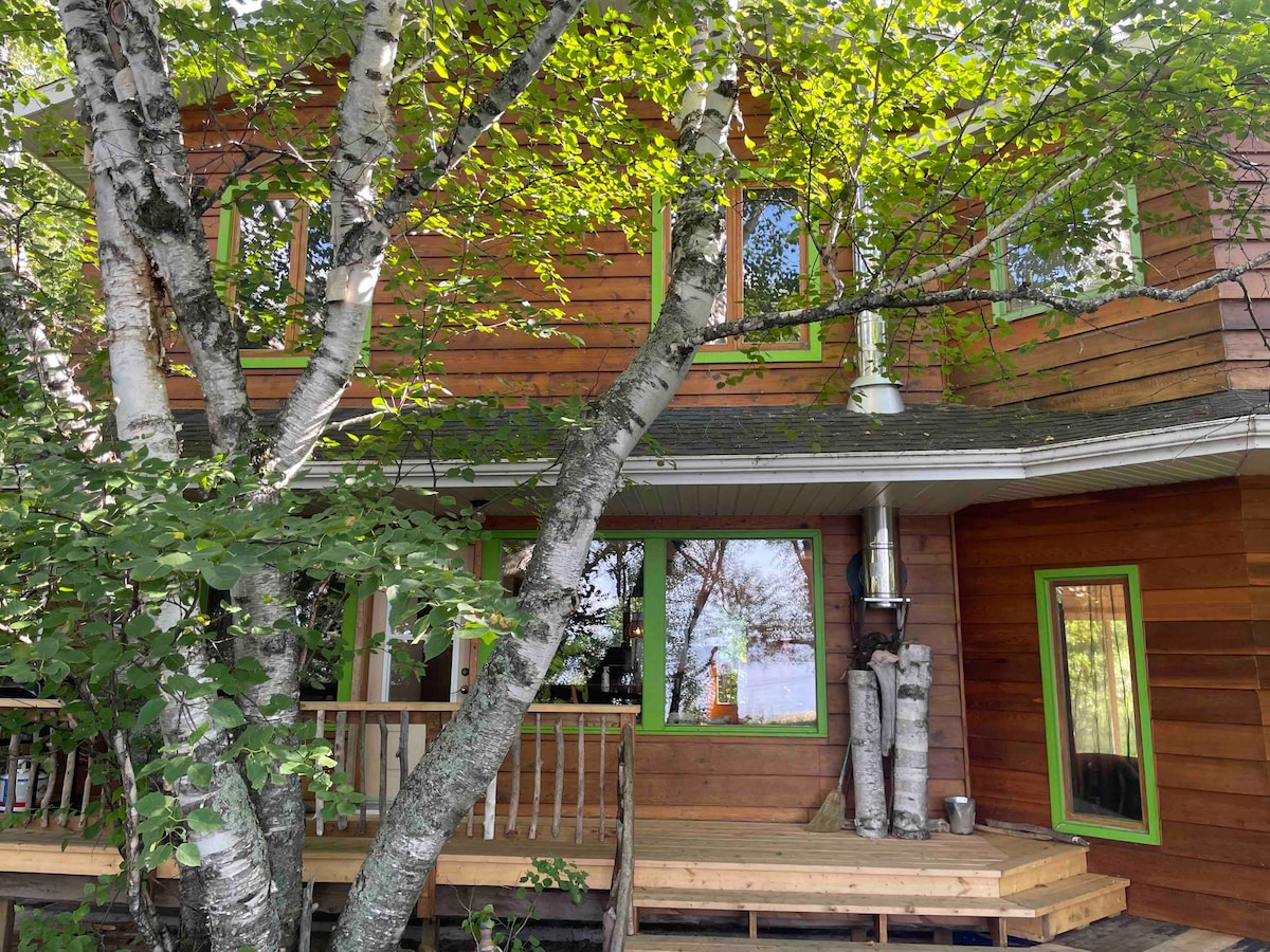The exterior of a wooden chalet is framed by lush greenery, with large windows reflecting the natural surroundings. A wooden porch is accessible from the entrance, featuring wooden railings and a cozy seating area. A visible chimney adds to the rustic charm.