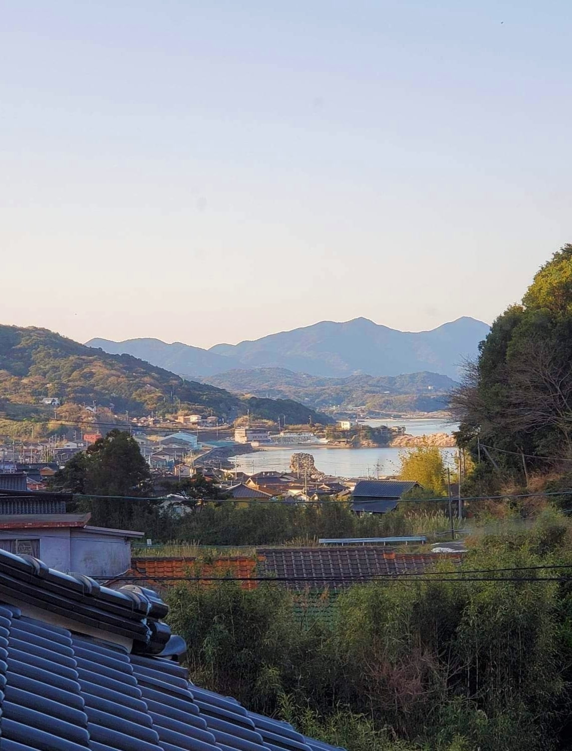 A serene landscape is depicted, featuring rolling mountains in the background and a tranquil body of water reflecting soft evening light. Traditional rooftops are visible in the foreground, creating a harmonious blend of nature and architecture.