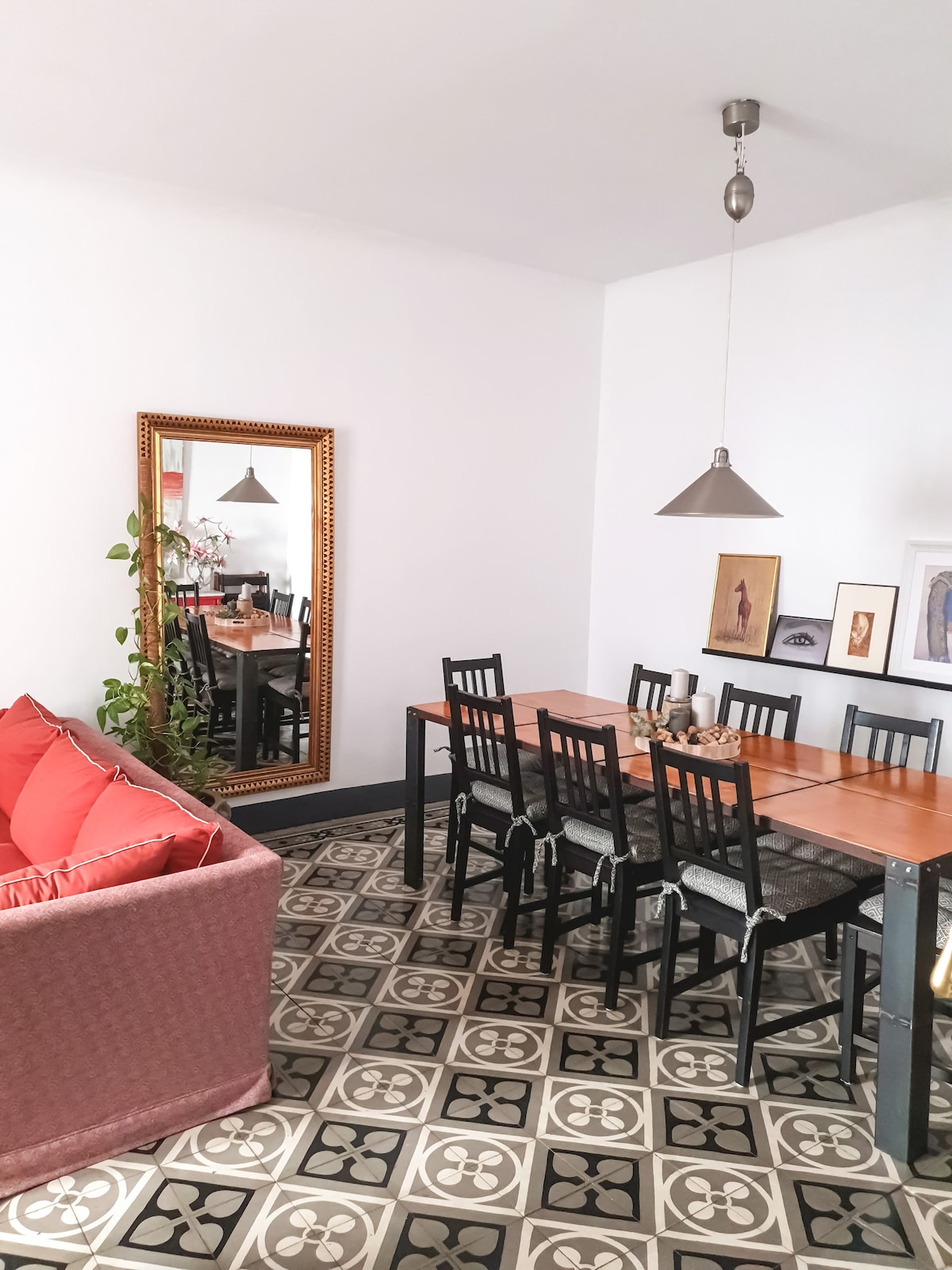 A dining area is visible, featuring a long wooden table surrounded by black chairs. A cozy seating space with red cushions is situated nearby. A large mirror reflects the light, while artwork is displayed on the walls, complementing the patterned tile flooring.