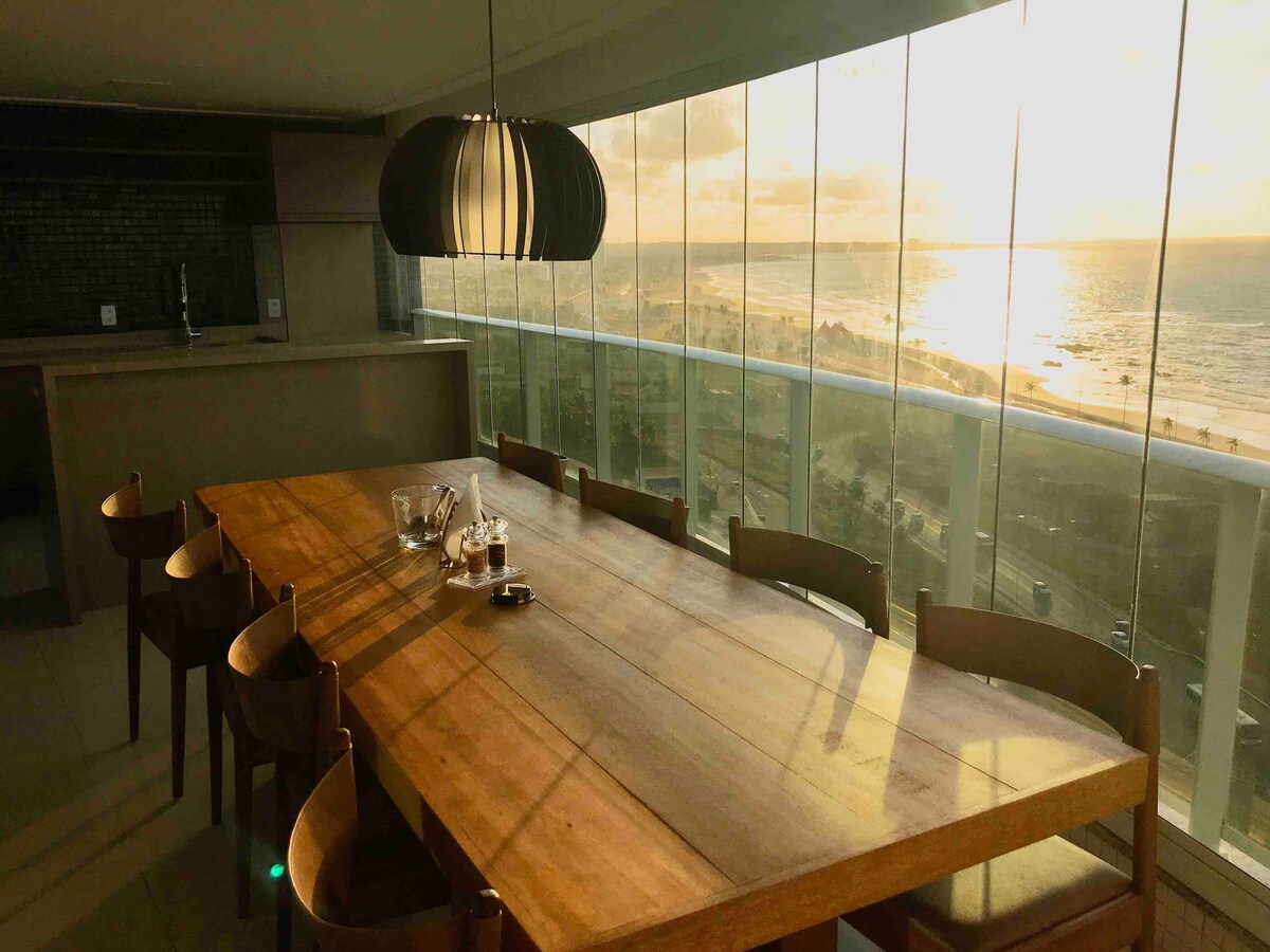 A large wooden dining table is positioned by floor-to-ceiling glass windows, offering a view of the ocean and coastline. Soft natural light streams in, highlighting the sleek design of the space. Surrounding chairs complement the table, creating a warm and inviting dining area.