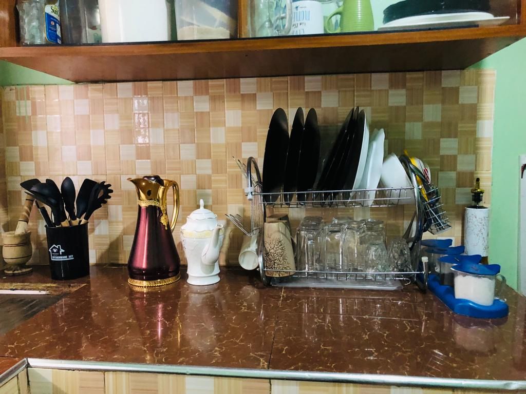 A kitchen countertop is populated with various utensils, including cooking tools and serving dishes. A dish rack holds neatly arranged plates and glasses, while decorative items are displayed alongside. The mosaic tile backsplash adds texture to the scene.