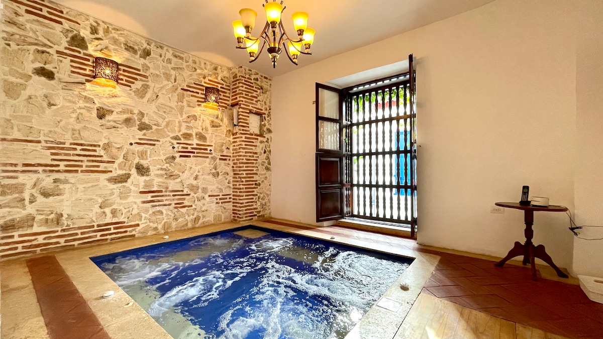 An indoor pool is set within a stone wall backdrop, illuminated by wall sconces. A large wooden door allows natural light to fill the space, while a small round table sits nearby. Soft ripples can be seen on the water's surface.