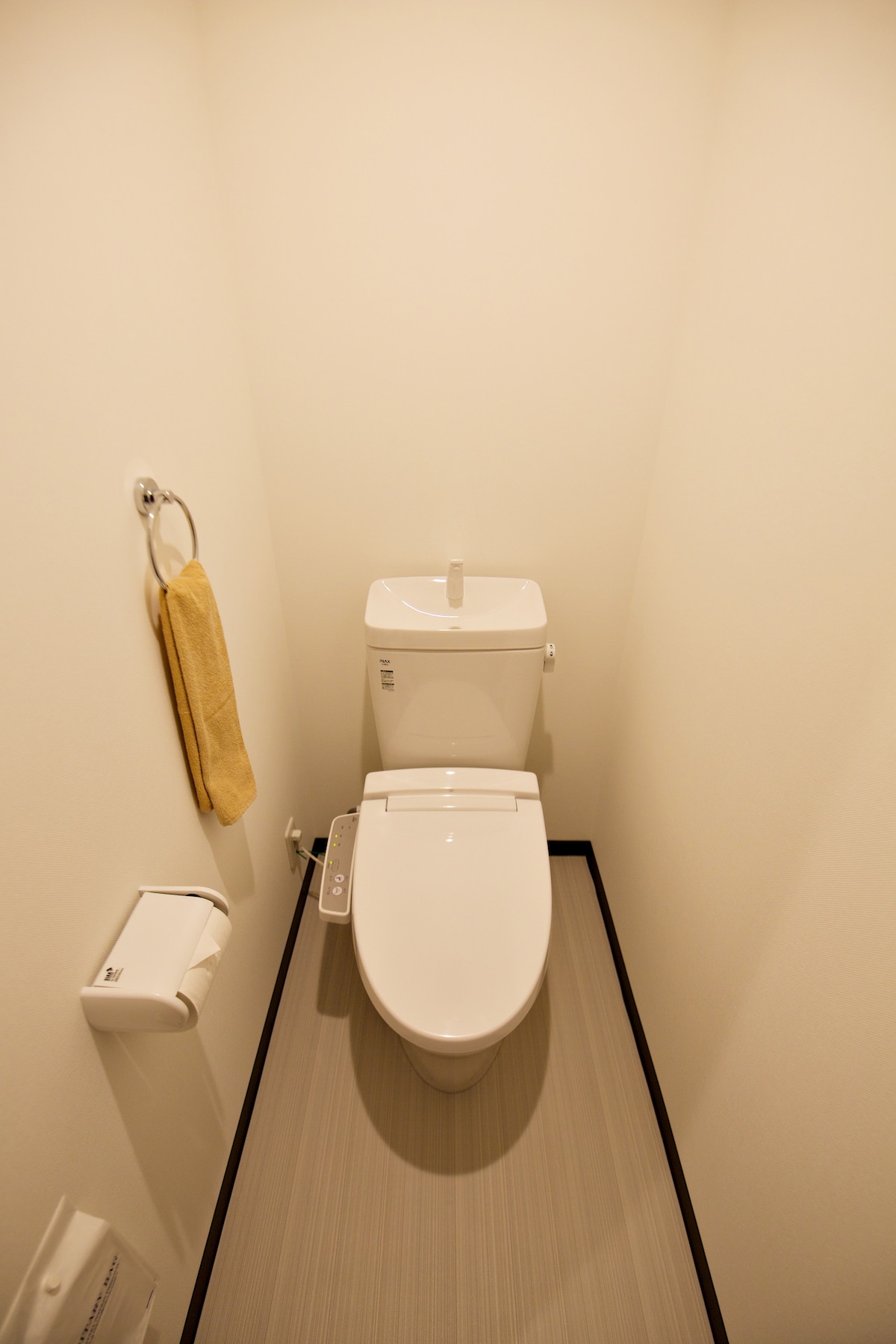 The bathroom features a modern toilet positioned against a smooth white wall. A roll of toilet paper and a towel hang nearby for convenience. The floor is covered in a light-colored material, contributing to a clean and minimalistic appearance.