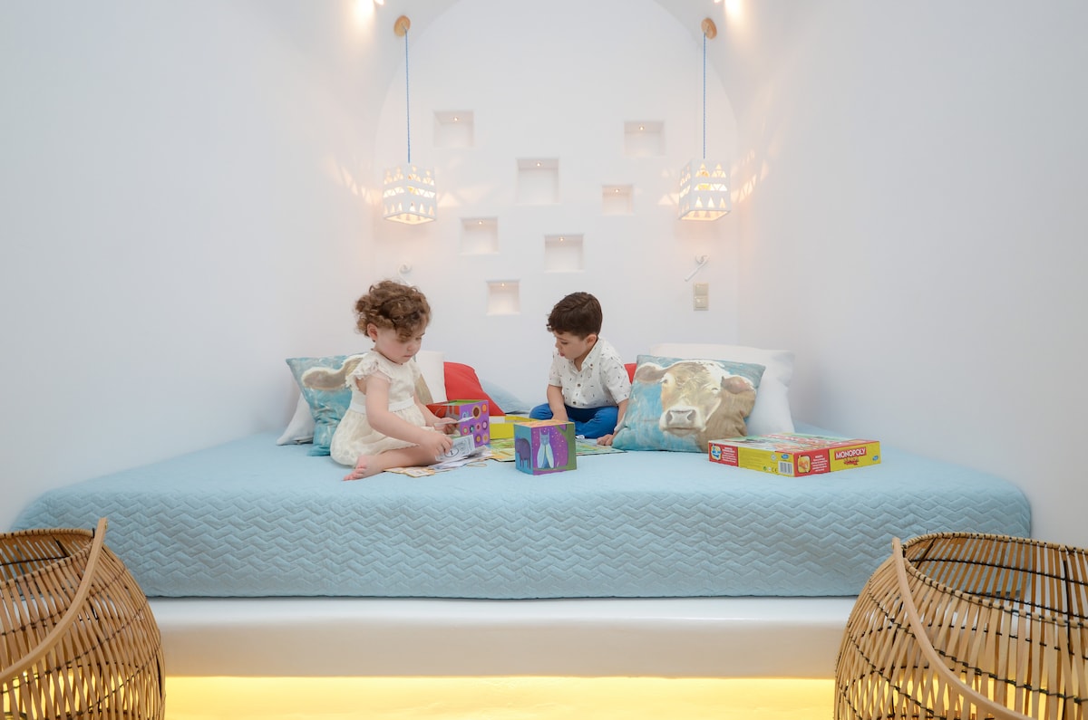 A traditional sleeping area is presented with a spacious bed adorned in a soft blue cover. Two colorful pillows provide comfort, while children engage with vibrant toys on the bed. Light fixtures above contribute to a bright atmosphere, complementing the minimalist decor.