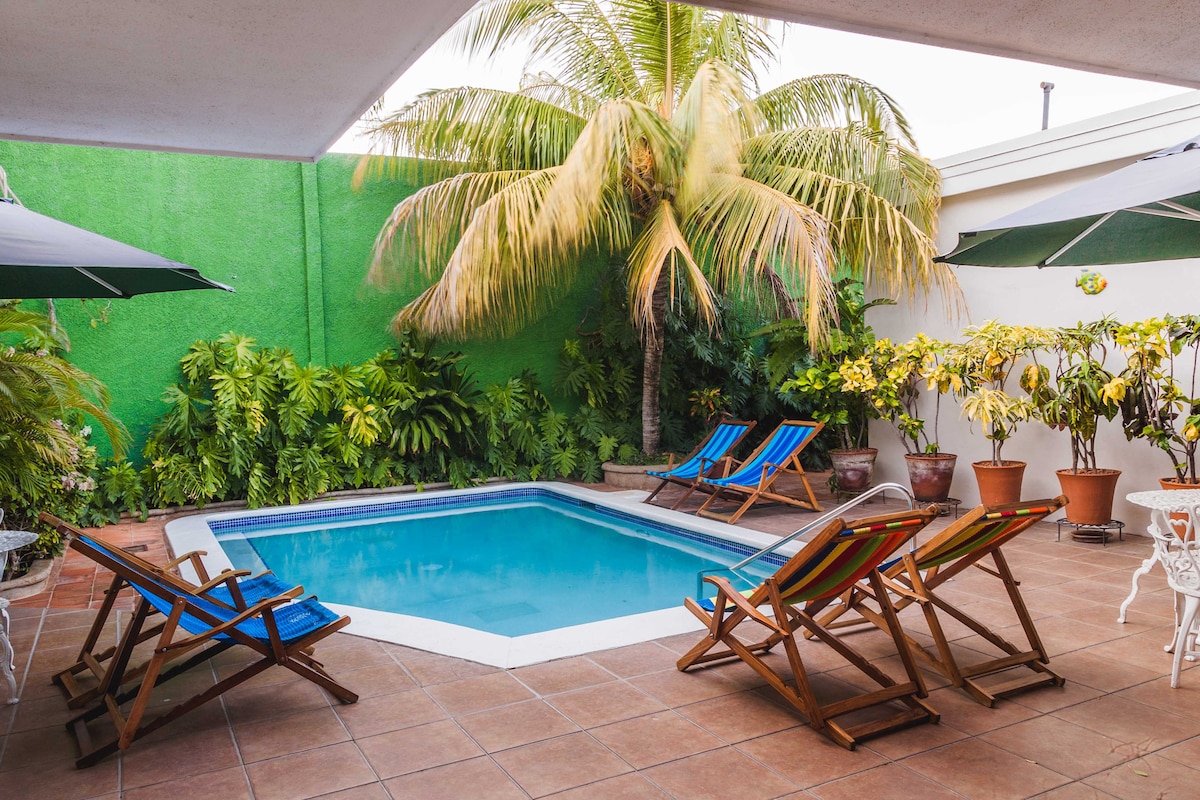 A private pool area is surrounded by lush greenery, featuring tropical plants and palm trees. Colorful lounge chairs in shades of blue and orange are arranged around the water, inviting relaxation. A tiled patio completes the outdoor space, offering a tranquil retreat.