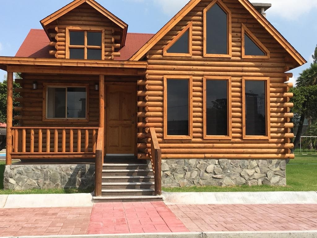 A wooden cabin is presented with a rustic design, featuring large windows that allow natural light to illuminate the interior. The entrance showcases a sturdy door framed by logs, and a stone foundation supports the structure. Steps lead up to the inviting porch.
