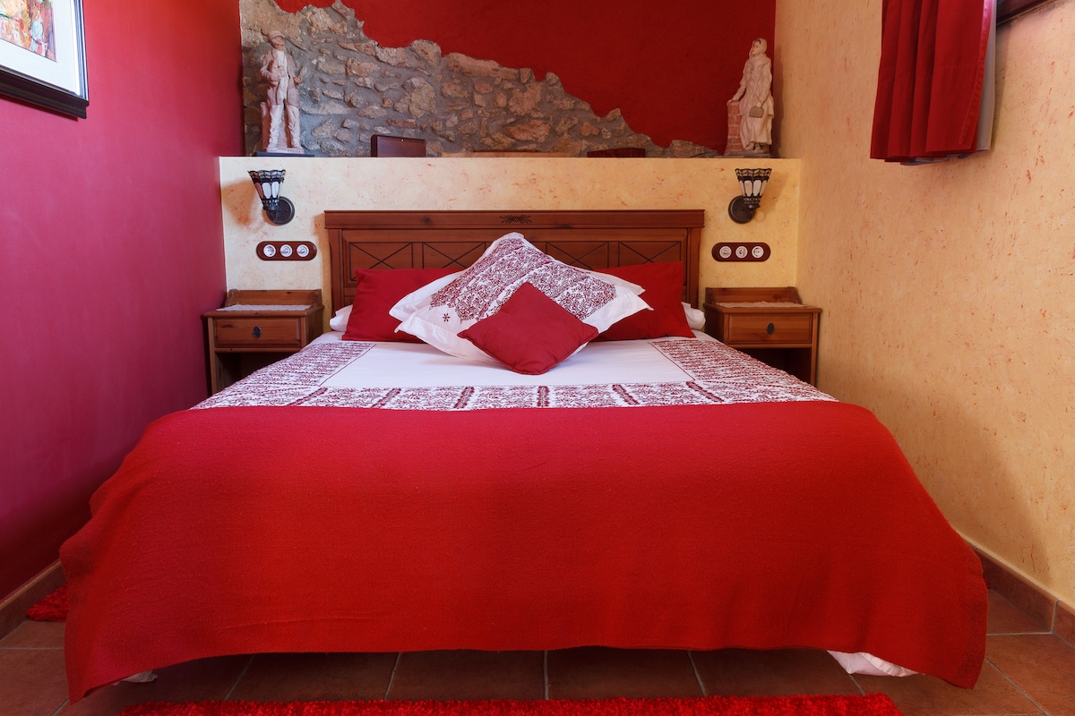 A spacious bed is dressed in rich red linens, complemented by decorative pillows. Flanking the bed are two wooden nightstands with stylish fixtures. The room features textured walls in warm tones, and a stone accent is visible in the background, enhancing the rustic charm.