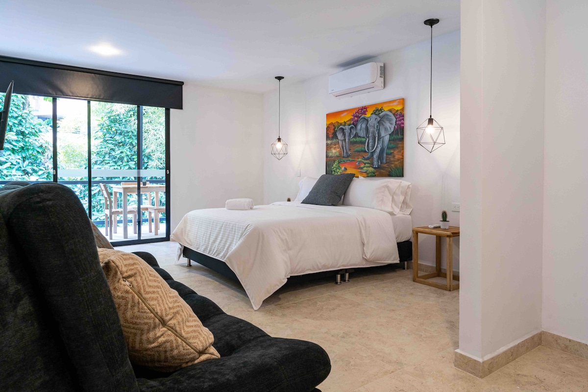 A spacious bedroom features a large bed with white linens, complemented by a decorative painting above. Natural light fills the room through a sliding glass door that opens to a small outdoor area with greenery. A cozy sitting area includes a dark, plush chair.
