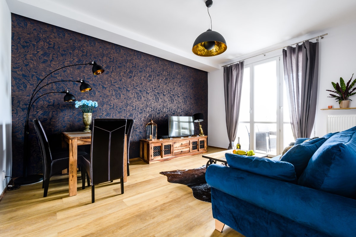 A spacious living area is featured, highlighted by a deep blue couch and a wooden dining table set for four. The two windows allow natural light, illuminated by stylish overhead lighting. Dark floral wallpaper provides an elegant backdrop, enhancing the modern aesthetic.