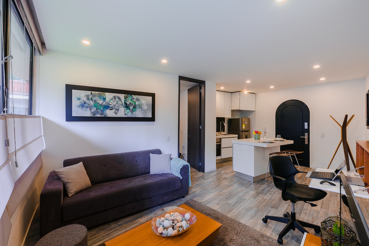 A modern living space features a dark sofa and a wooden coffee table adorned with decorative elements. A sleek desk and chair are positioned near large windows, enhancing natural light. An open kitchen area is visible in the background, showcasing contemporary appliances.