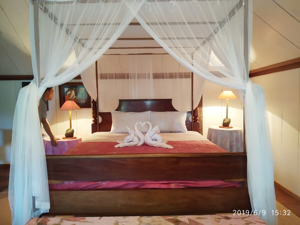 A spacious bungalow bedroom features a wooden bed draped with sheer white curtains. Two elegantly arranged towel swans sit on the covers, complemented by bedside lamps on either side. Soft light falls on the pink and neutral color tones of the bedding and surrounding decor.