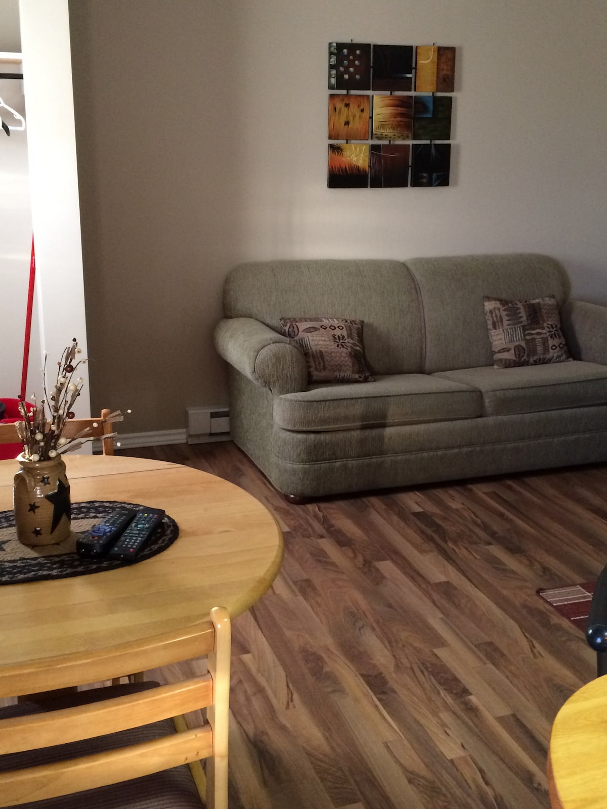 A light-filled living space features a green sofa adorned with patterned cushions. A round wooden dining table with light-colored chairs is positioned nearby. A decorative vase with twigs and a remote control can be seen on a circular placemat, enhancing the room's comfort.