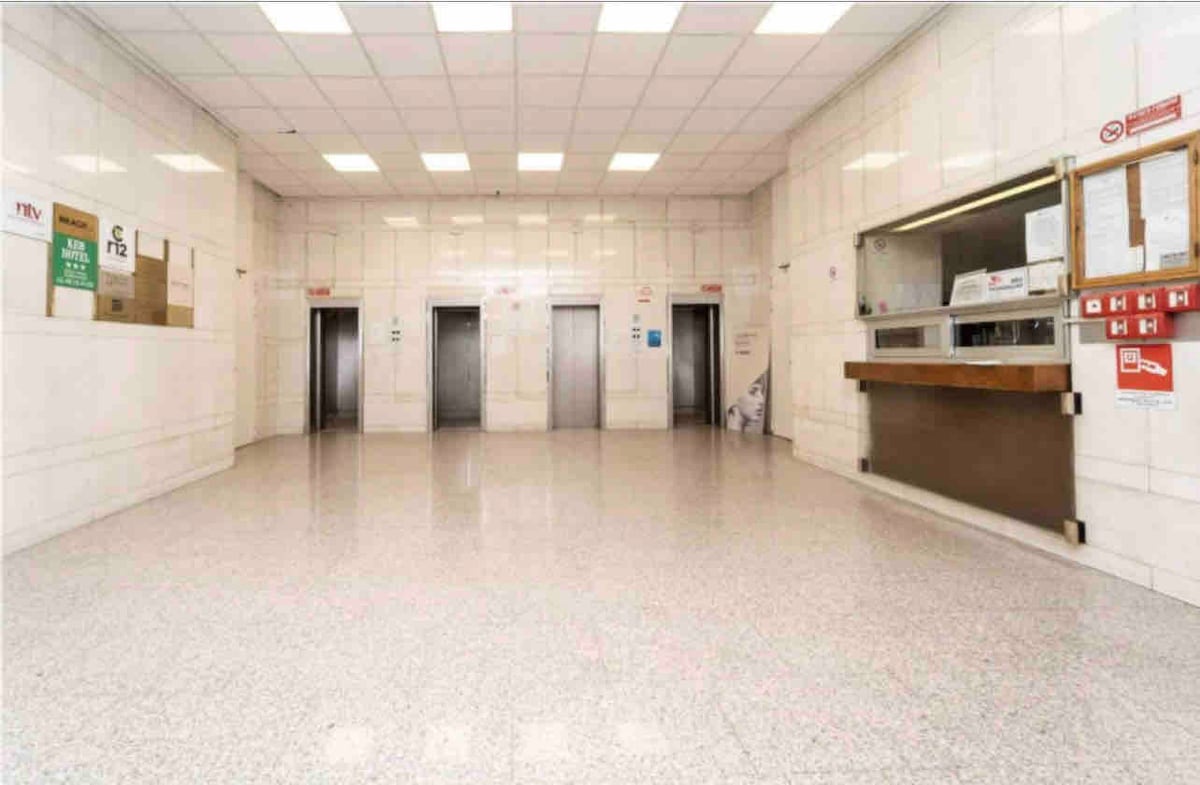 A spacious lobby features shiny tiled floors and brightly lit ceilings. Four elevator doors are visible on one side, and a reception area with a wooden counter is located on the adjacent wall. Clean and well-maintained, the space offers easy access to building amenities.