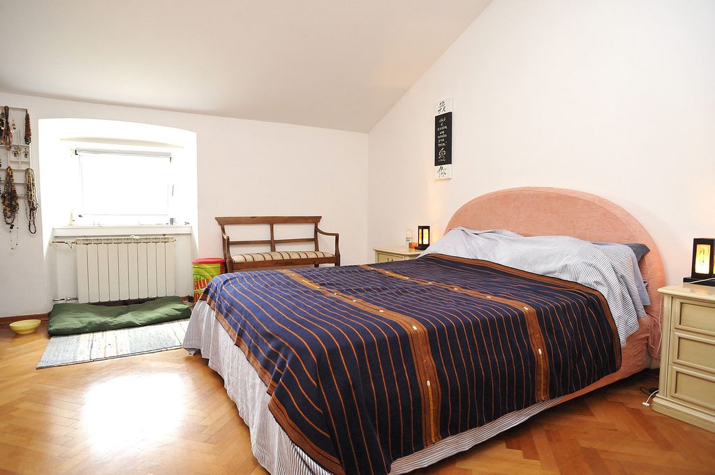 A spacious bedroom features a large bed with a striped blanket. Two bedside tables are positioned beside the bed, and a wooden chair is located at the foot. Natural light enters through a window, providing a bright atmosphere.