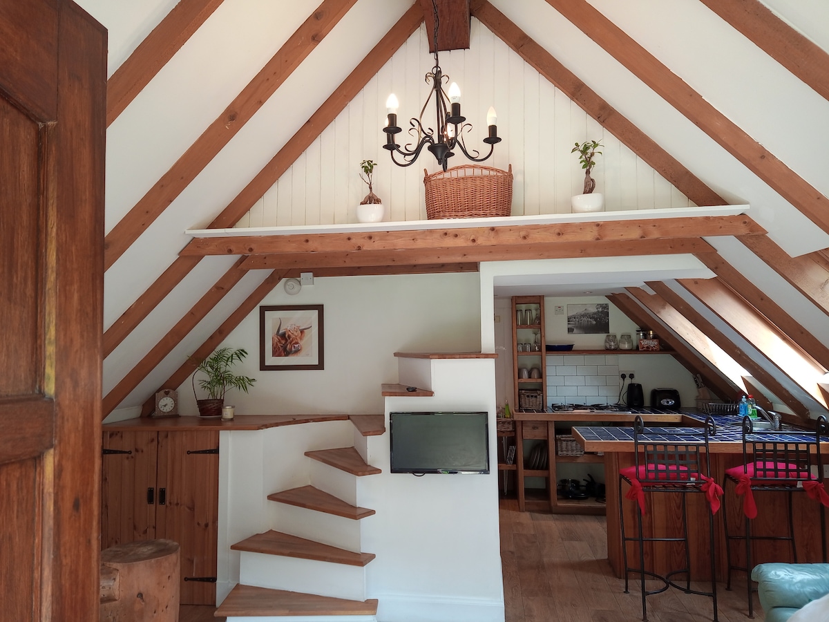 A ceiling with exposed wooden beams creates a charming atmosphere. A chandelier hangs above an open space that includes a dining area with seating. Stairs on the left lead to an upper level, while a small television is mounted on the wall nearby.