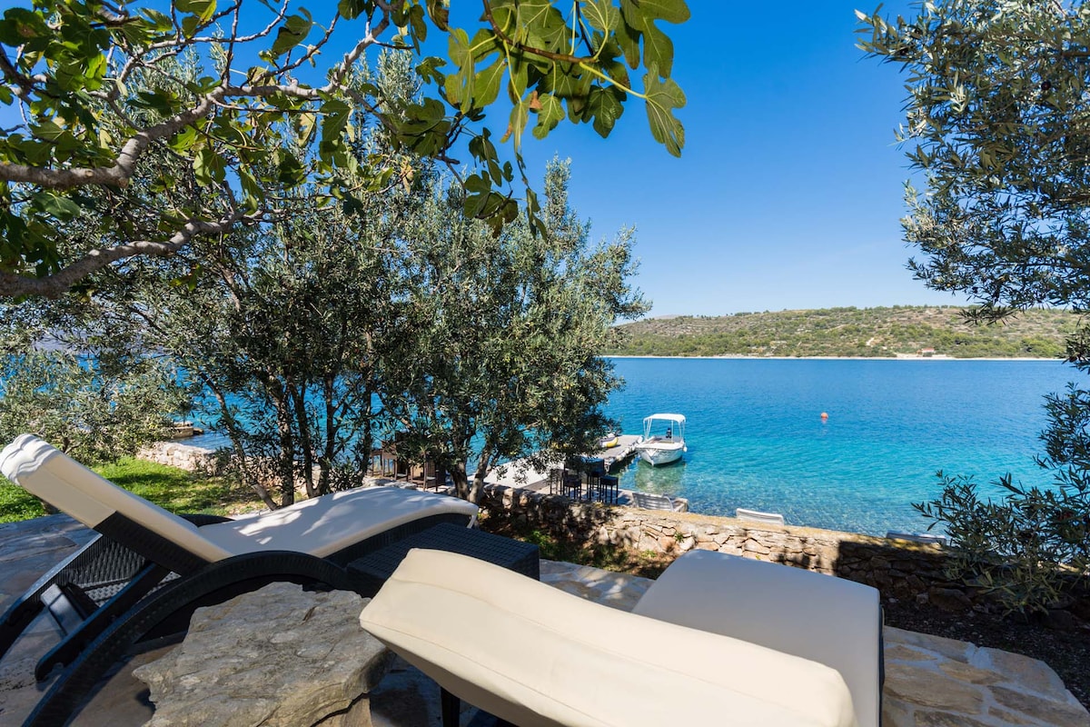 Two sunbeds are positioned on a stone terrace, overlooking azure waters. Olive trees provide shade, and a small boat is visible in the distance. The tranquil scene captures the natural beauty and inviting atmosphere of the private beach area.