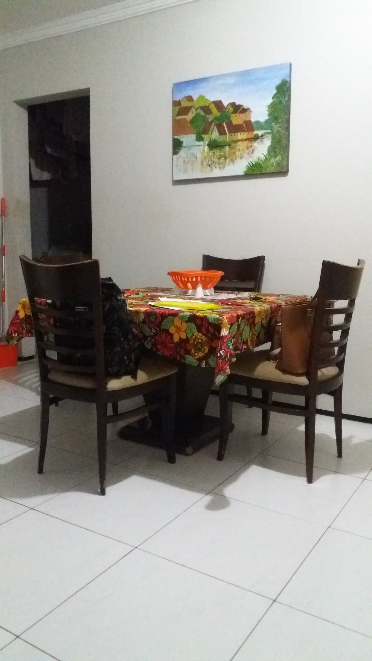 A dining area is presented with a square table adorned with a colorful patterned tablecloth. Four wooden chairs are positioned around the table. A fruit bowl sits at the center, and a piece of artwork featuring a rural scene is displayed on the wall.