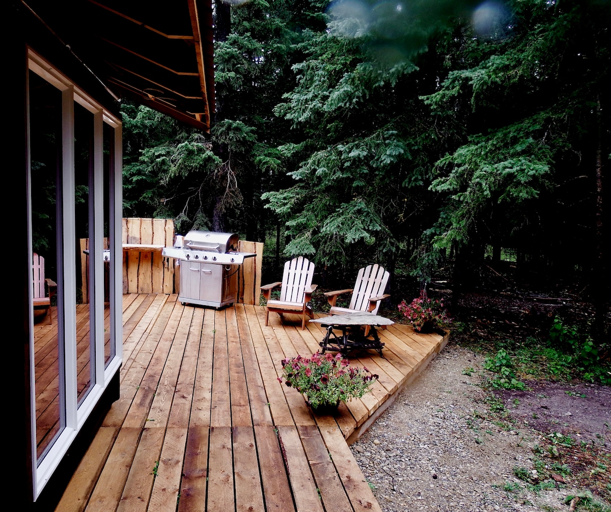 A spacious wooden deck features two Adirondack chairs and a propane BBQ. Surrounding greenery adds a natural backdrop, while planters with colorful flowers enhance the outdoor space. Access to the cabin is visible through large glass doors.