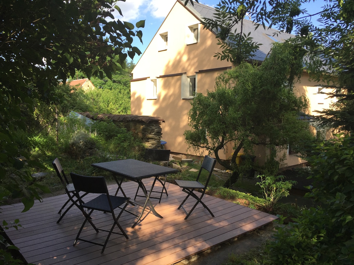 A private terrace is surrounded by greenery, featuring a table and four chairs. The pale exterior of the house is visible in the background, framed by trees and shrubs, creating a serene outdoor setting.