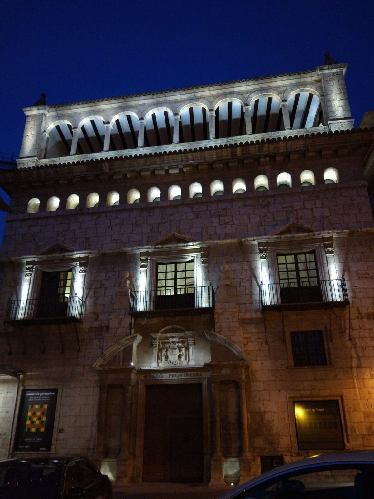 A historic building is illuminated at night, showcasing its architectural features. The stone facade is complemented by ornate balconies and large windows that reflect the ambient light. A patterned detail is visible on the ground, adding to the character of the entrance.