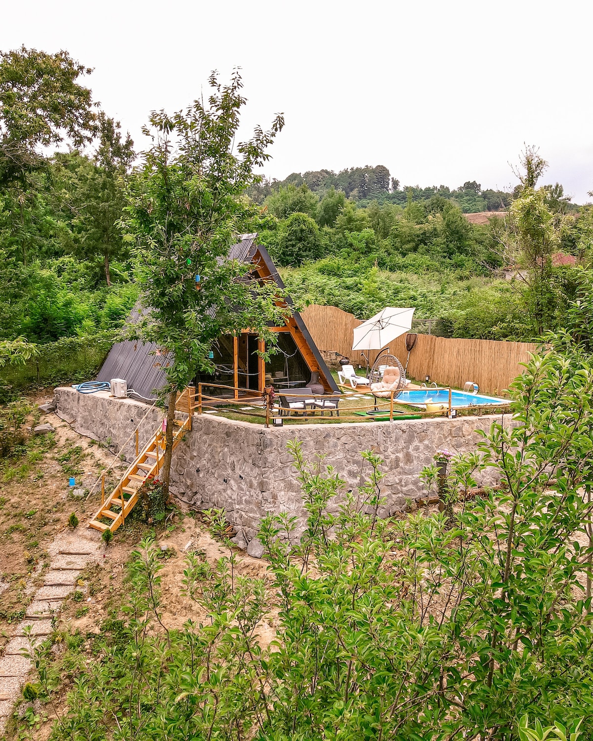 A unique triangular-shaped bungalow is nestled among lush greenery, surrounded by a stone wall. A wooden staircase leads to the entrance, while a spacious outdoor area with a pool can be seen, complete with a sunshade and comfortable seating.