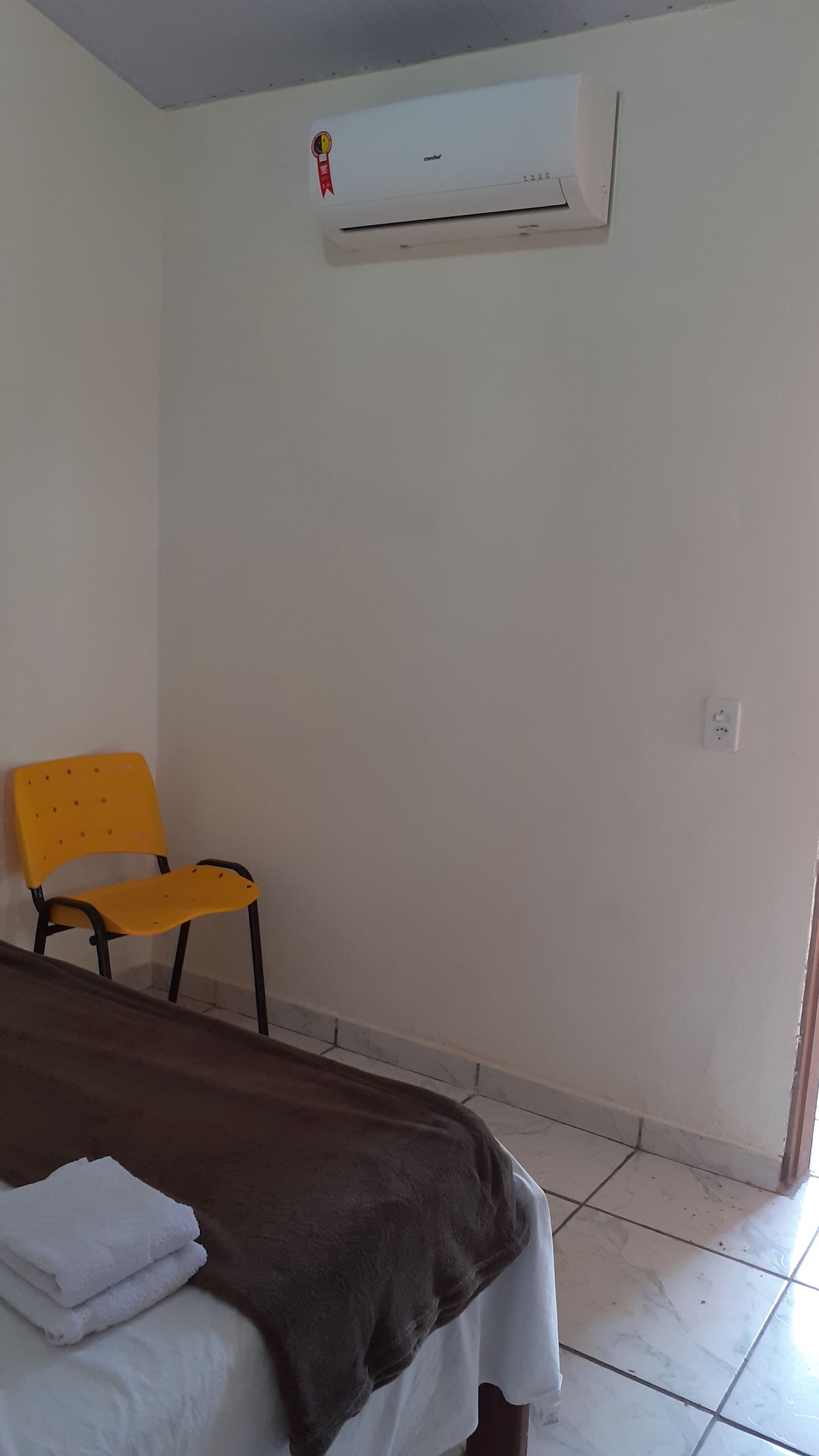A simple room features a spacious bed covered with a dark blanket. A yellow chair is positioned near the wall, and an air conditioning unit is installed above. Soft light enters from a nearby doorway, illuminating the tiled floor.