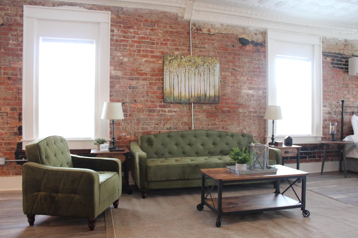 A cozy living area features a green upholstered sofa and an accent chair, complemented by a wooden coffee table. Exposed brick walls add character, while large windows provide natural light. A piece of artwork depicting trees enhances the space, with table lamps adding functional lighting.