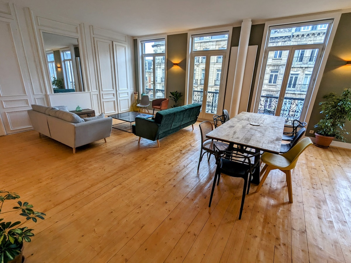 An open living space features large windows offering natural light and views of Haussmannian buildings. Two sofas, one in gray and one in green, are positioned alongside a wooden dining table surrounded by various chairs. The wooden floor enhances the room's brightness.