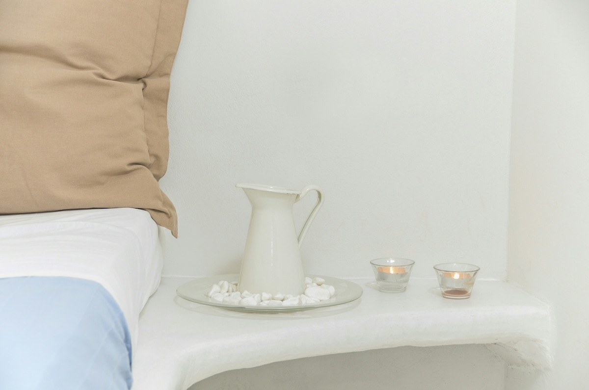 A tranquil bedside display features a white ceramic pitcher resting on a circular tray with small pebbles. Two glass candles are positioned nearby, creating a calming ambiance in a serene, neutral-toned environment.