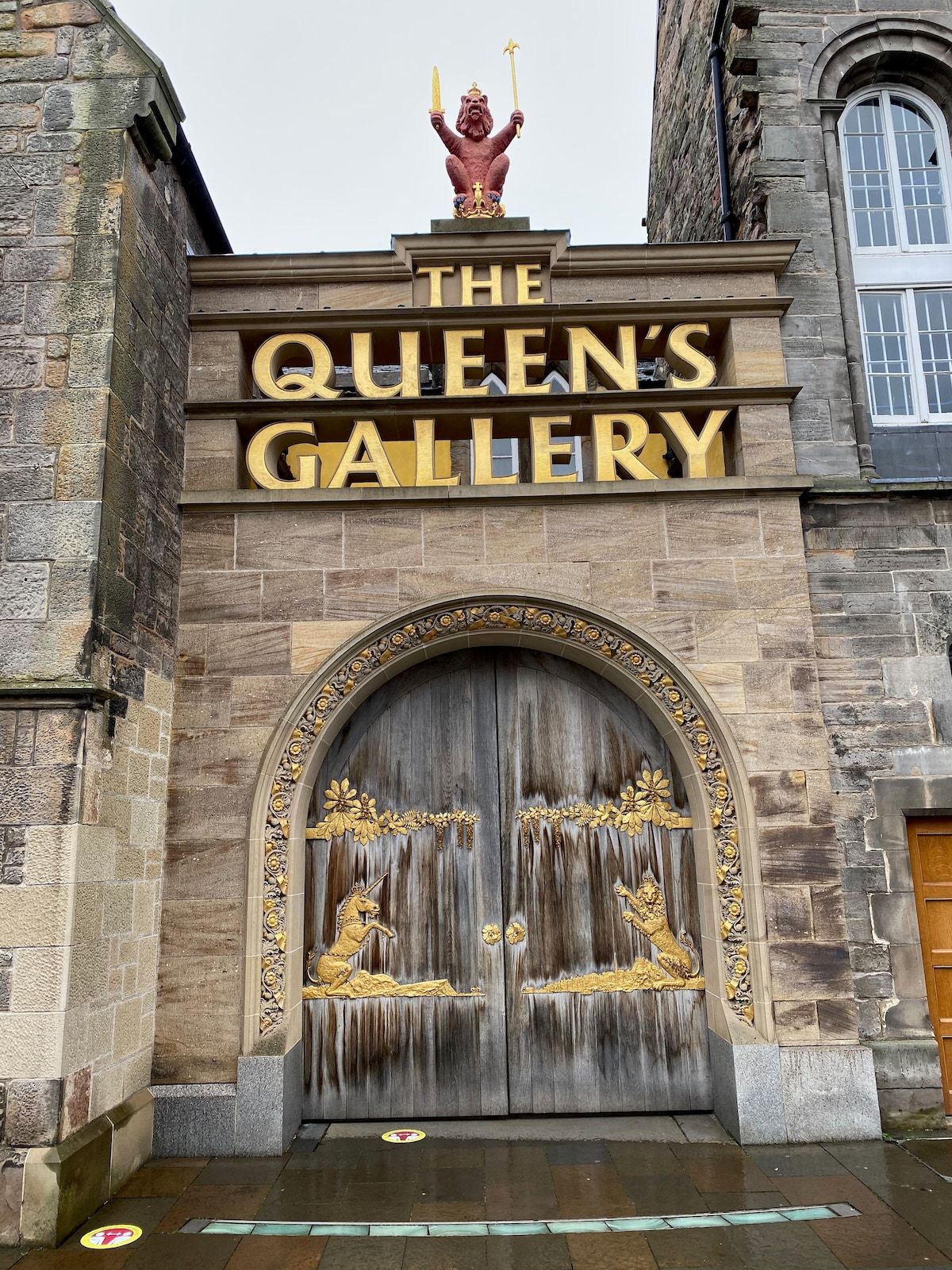 The entrance to The Queen's Gallery is showcased, featuring ornate wooden doors with intricate carvings of animals. A prominent golden sign displays the name of the gallery above the entrance, while a decorative figure holds a staff atop the building.