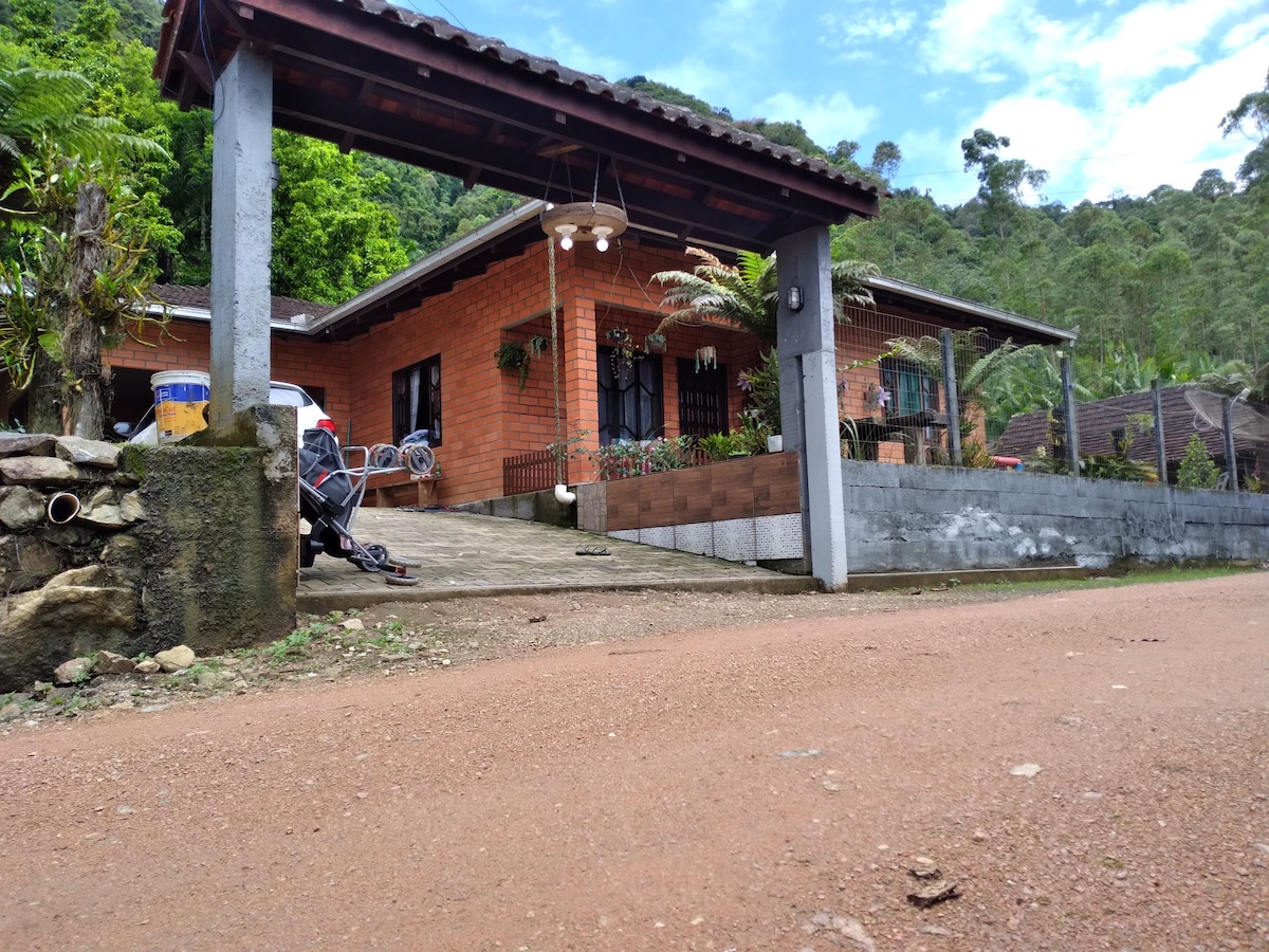 The entrance of a charming brick house is visible, framed by lush greenery and a serene mountain backdrop. A gravel path leads to the house, which features large windows and a welcoming porch. Decorative elements include hanging lights and a stone wall.