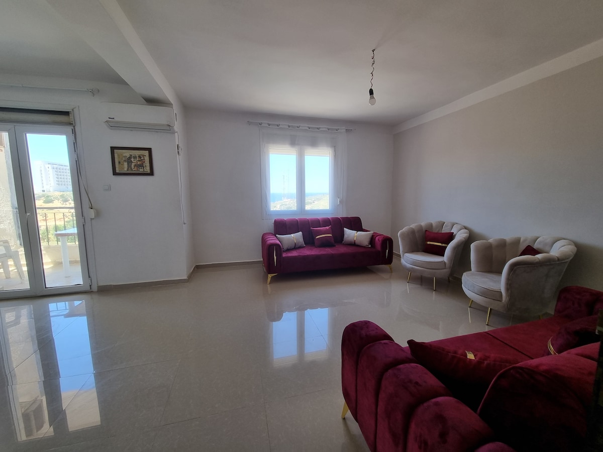 A spacious living area features plush maroon sofas and cream armchairs arranged thoughtfully. Natural light floods the room through large glass doors that open to a balcony, enhancing the bright atmosphere. A sleek floor reflects the light, contributing to the modern aesthetic.