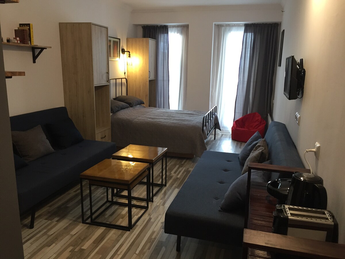 A studio space is furnished with a double bed and two expandable sofas, accompanied by a set of wooden coffee tables. Natural light filters through large windows draped with dark curtains. A kitchenette area includes a coffee maker and a small toaster.
