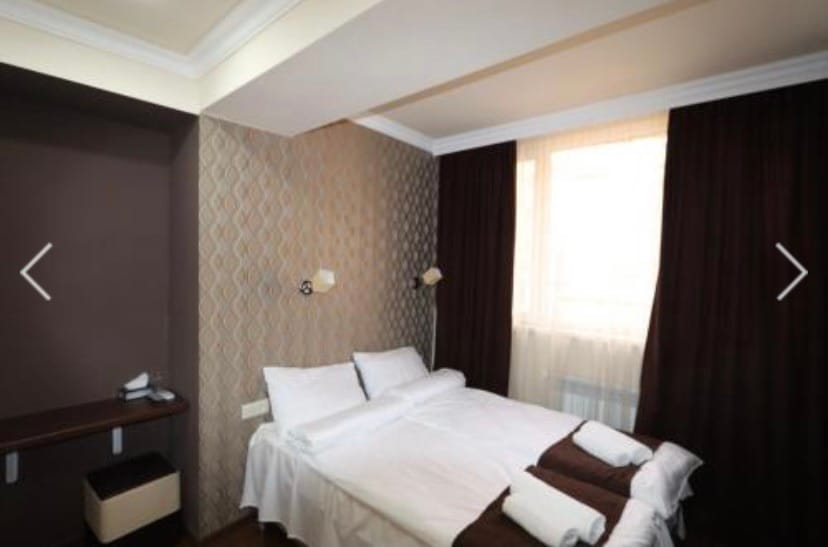 A cozy sleeping area is presented with a spacious bed featuring crisp white linens and dark brown accents. Soft lighting is provided by wall-mounted lamps. Neutral-toned walls complement the dark curtains covering the large window, allowing natural light to fill the room.